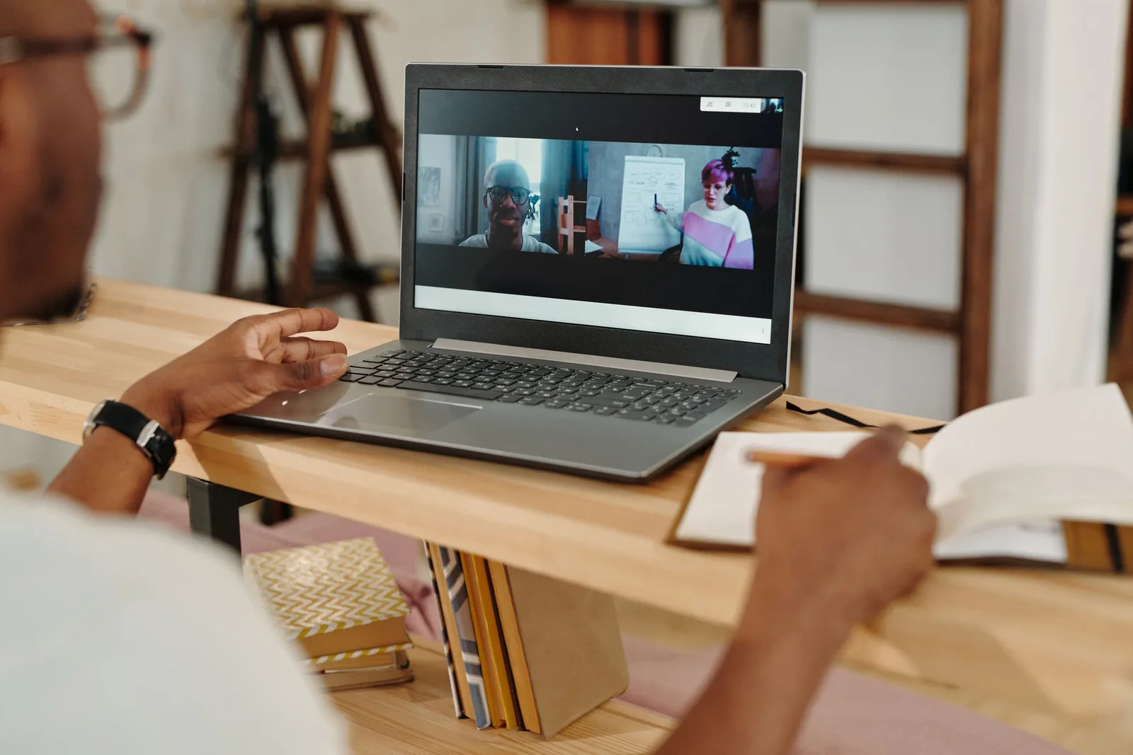Laptop-এ video meeting চলার সময় notebook-এ notes লিখছেন একজন ব্যক্তি