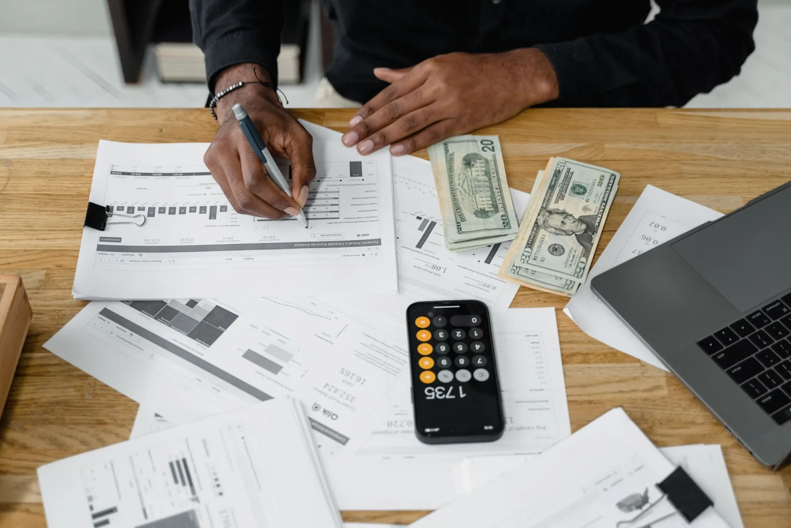 Laptop, calculator ও financial reports-এর পাশে payout হিসাব মিলিয়ে লিখছেন একজন ব্যক্তি